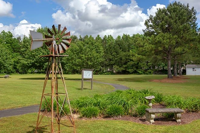A rustic windmill stands in a lush garden with vibrant greenery and a backdrop of trees under a cloudy sky.