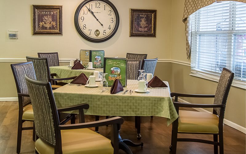 A cozy dining room with neatly set tables, featuring a large wall clock and comfortable seating.