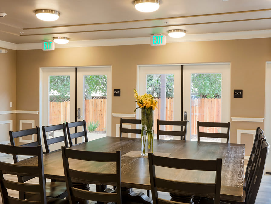 A dining room with a large table, chairs, and a vase of flowers near the windows.