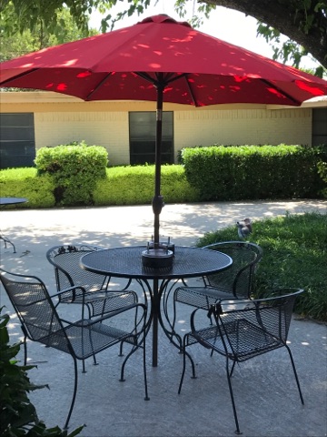 A cozy outdoor dining setup featuring a round table with four chairs and a large red umbrella, surrounded by lush greenery.