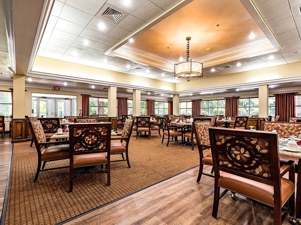 A spacious and elegantly designed dining room with ornate chairs and tables, featuring warm lighting and a carpeted floor.