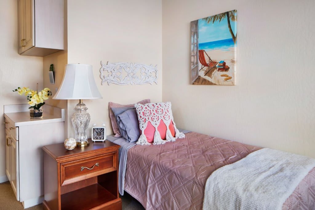 A cozy bedroom featuring a single bed with decorative pillows, a nightstand with a lamp, and beach-themed wall art.