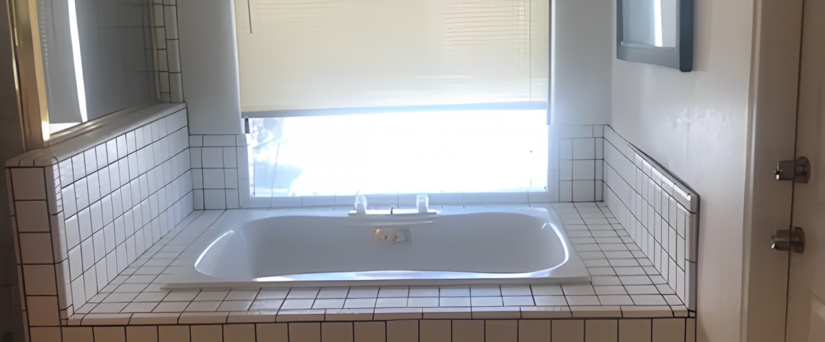 A sunlit bathroom featuring a white bathtub beneath a window with blinds, surrounded by tiled walls.