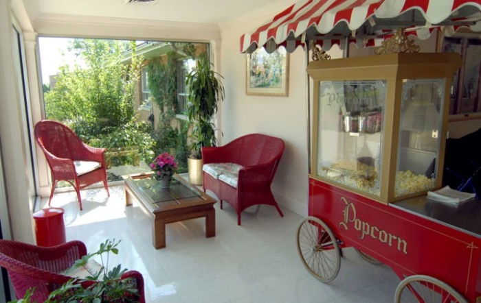A cozy indoor space featuring a popcorn machine, red chairs, a coffee table, and lush plants with a garden view.