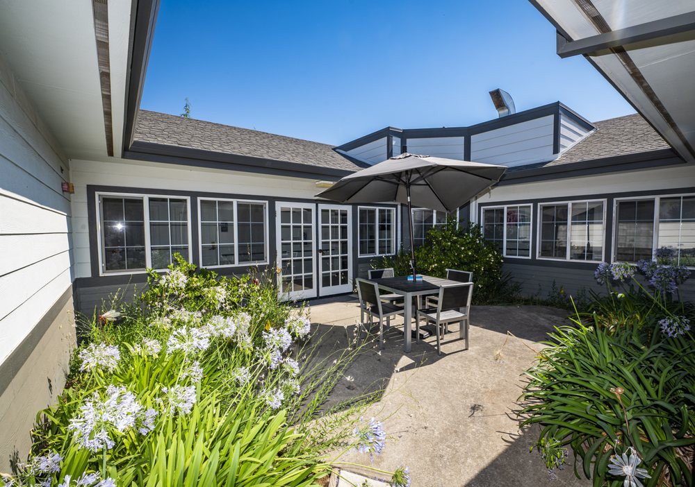 A cozy outdoor patio with a table, chairs, and an umbrella, surrounded by a garden and large windows of the house.