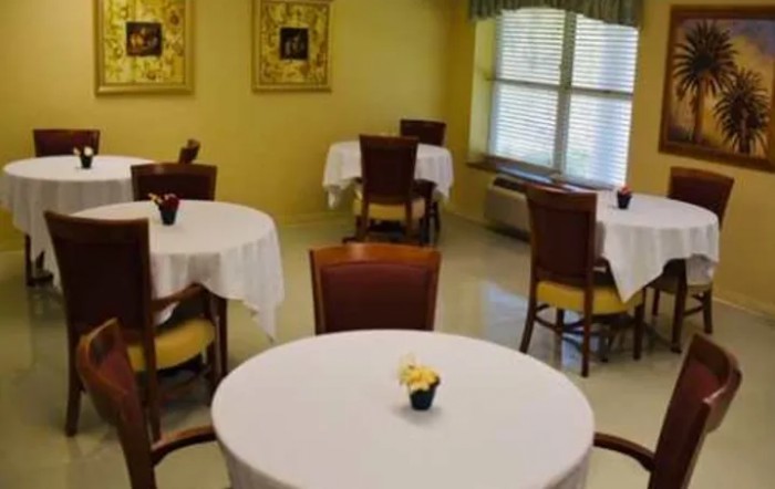 A quiet dining room with round tables and chairs arranged neatly, featuring white tablecloths and a window allowing in natural light.