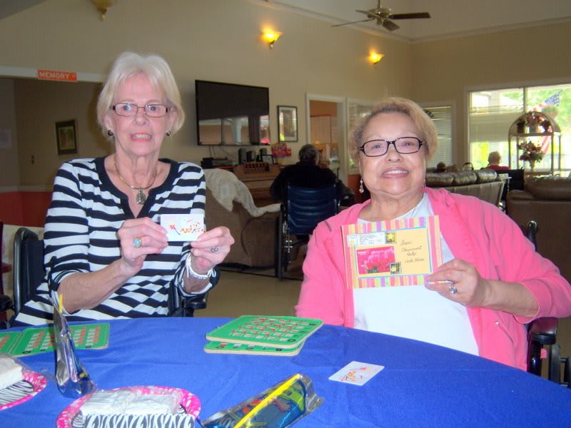 Two women smiling and holding cards while seated at a table indoors.