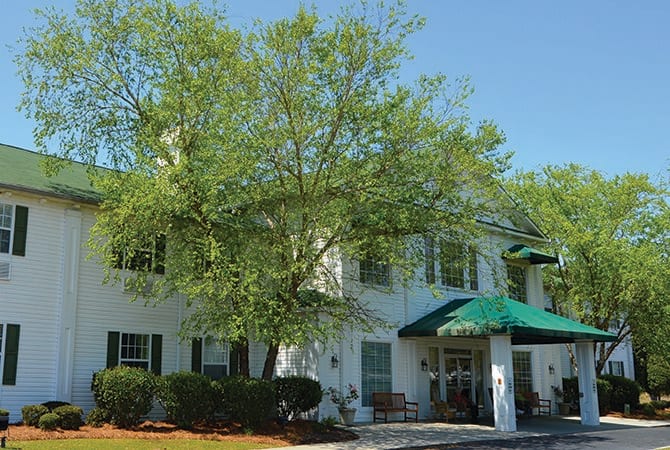 A sunny view of a building with white walls, surrounded by green trees and a clear blue sky.