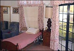 A cozy bedroom features a single bed with a pink cover, a lamp on a wooden nightstand, and floral curtains framing the window.