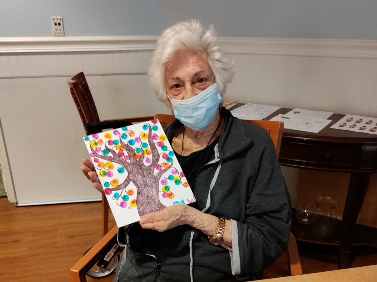 An elderly woman wearing a face mask holds up her colorful tree drawing while seated indoors.