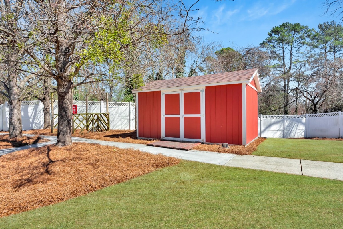A red garden shed in a backyard with a lawn, tree, and white fence.