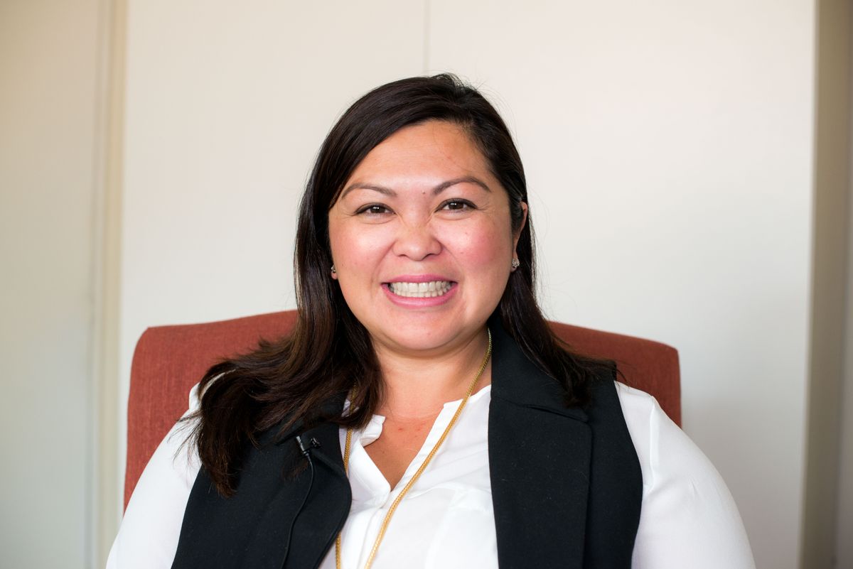 A smiling woman is seated indoors, wearing a black vest over a white shirt.