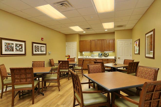 A well-lit dining area with several tables and chairs, adjacent to a kitchenette.