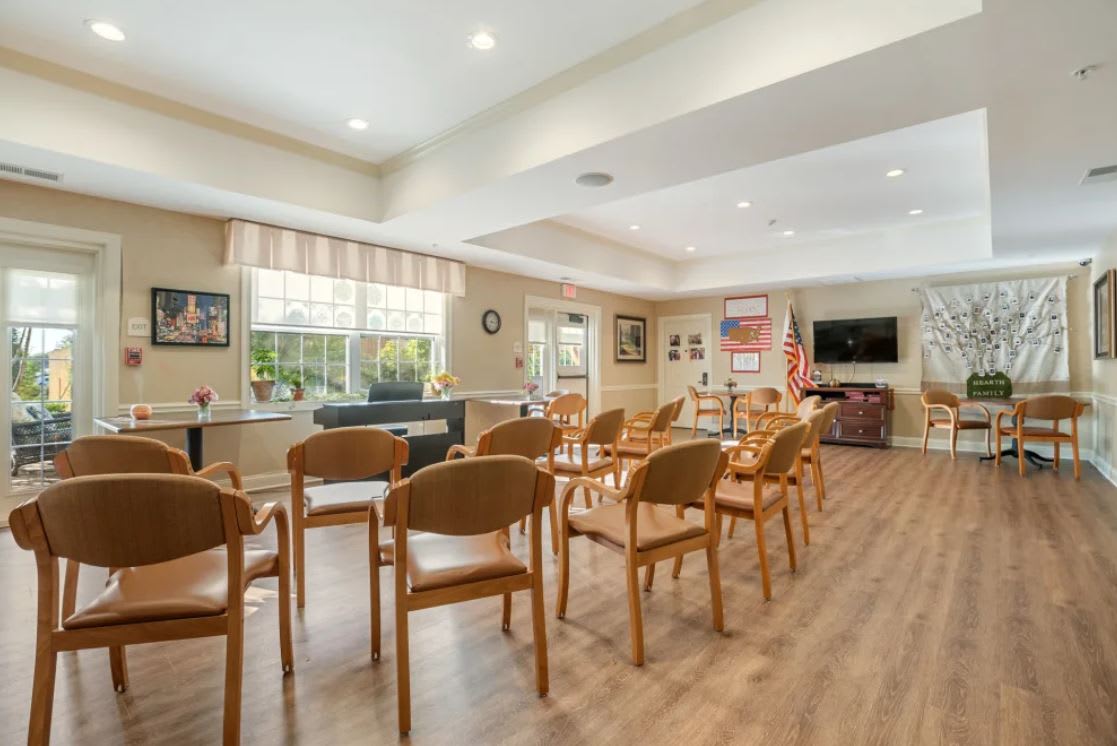 A well-lit meeting room with wooden chairs arranged facing a television and American flags, featuring large windows and a wooden floor.
