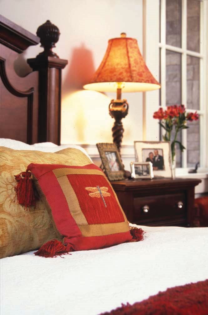 A cozy bedroom scene with decorative pillows, a bedside lamp, and a nightstand adorned with flowers and framed photos.