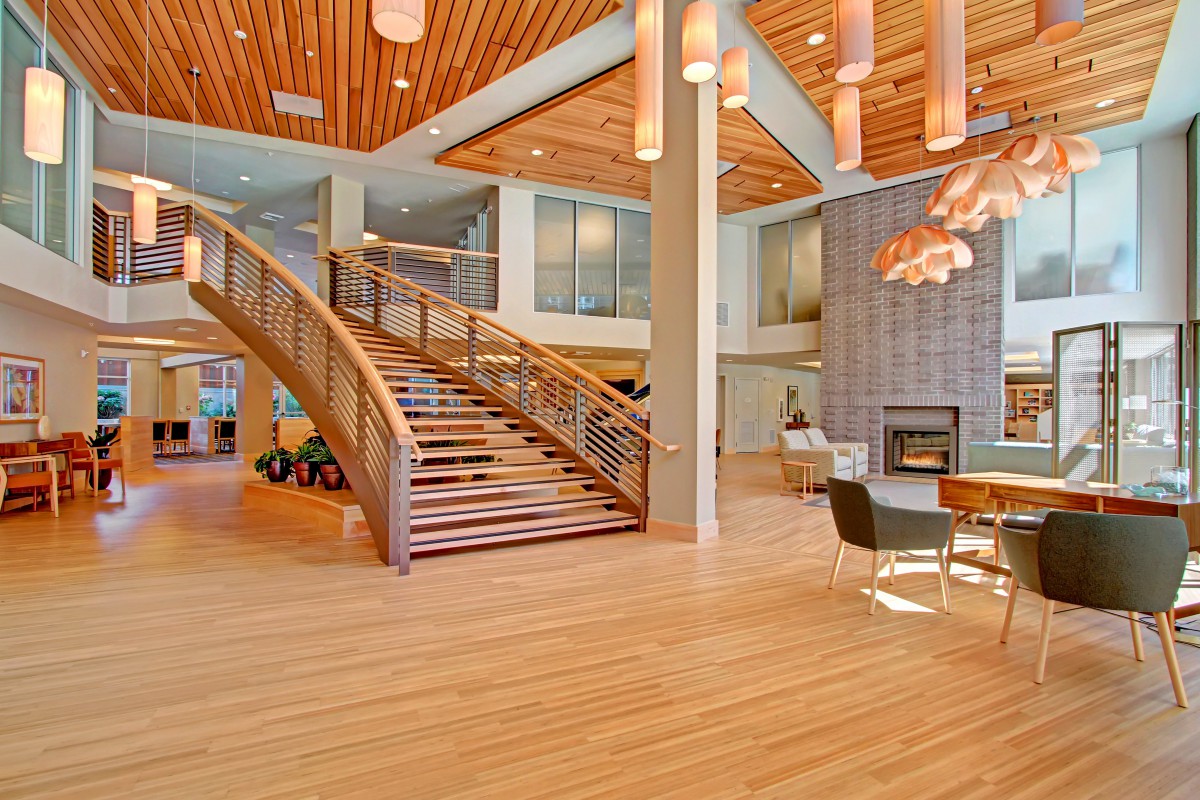 A spacious and modern lobby with wooden flooring, a grand staircase, stylish lighting fixtures, and comfortable seating around a fireplace.