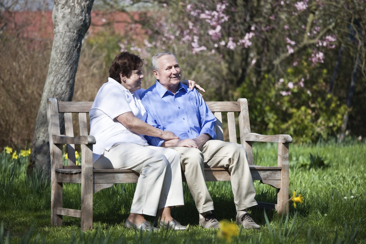 An elderly couple sitting together on a park bench amidst blooming trees.