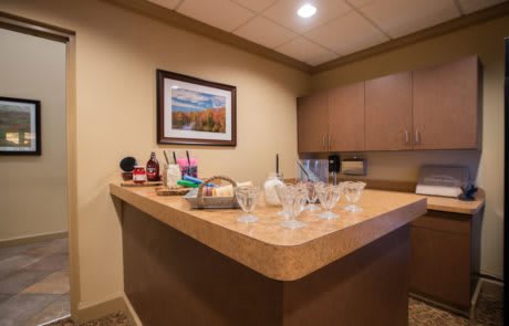 A small bar area with a countertop, assorted bottles, and glasses, set against a warm interior with cabinets and wall art.