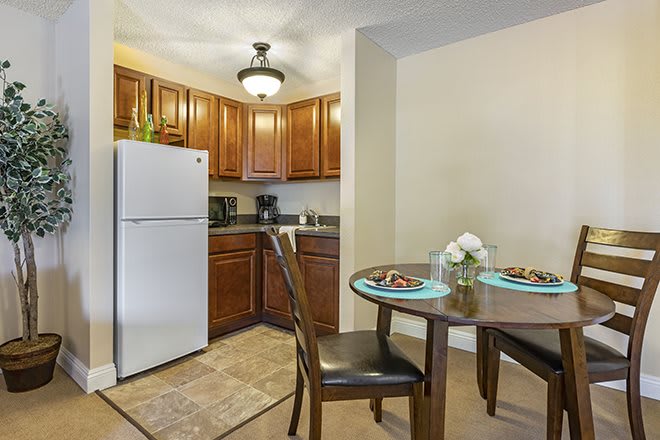 A cozy kitchen and dining area featuring wooden cabinets, a refrigerator, and a small dining table set for two with decorative flowers.