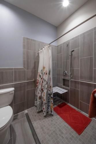 A modern bathroom with accessible features, including a tiled walk-in shower, shower curtain, grab bars, and a red rug.