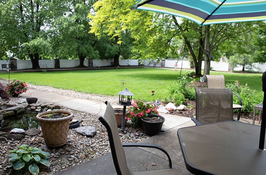A peaceful backyard patio with chairs, a table under an umbrella, and a well-maintained lawn surrounded by trees.