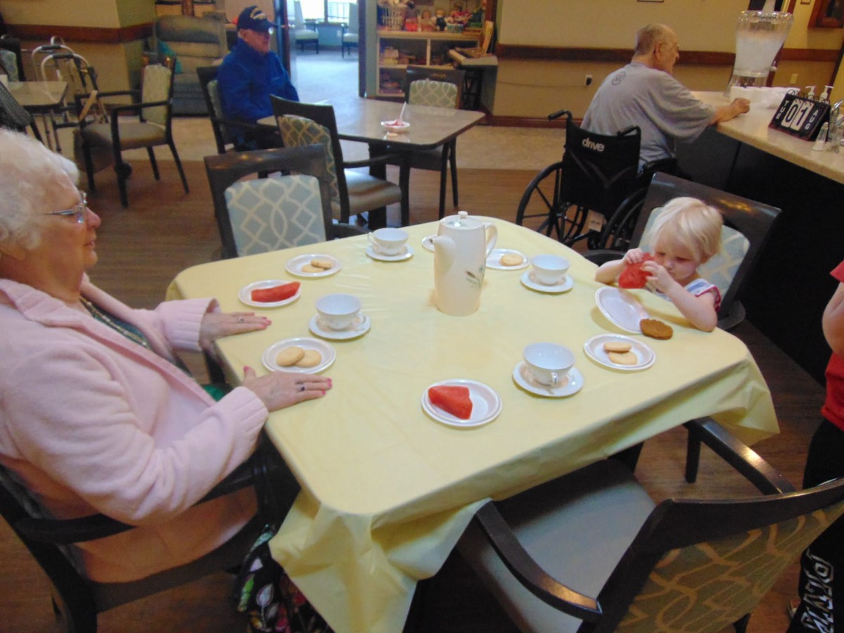 An elderly person and a child sit at a dining table set with tea cups and cookies in a communal setting.