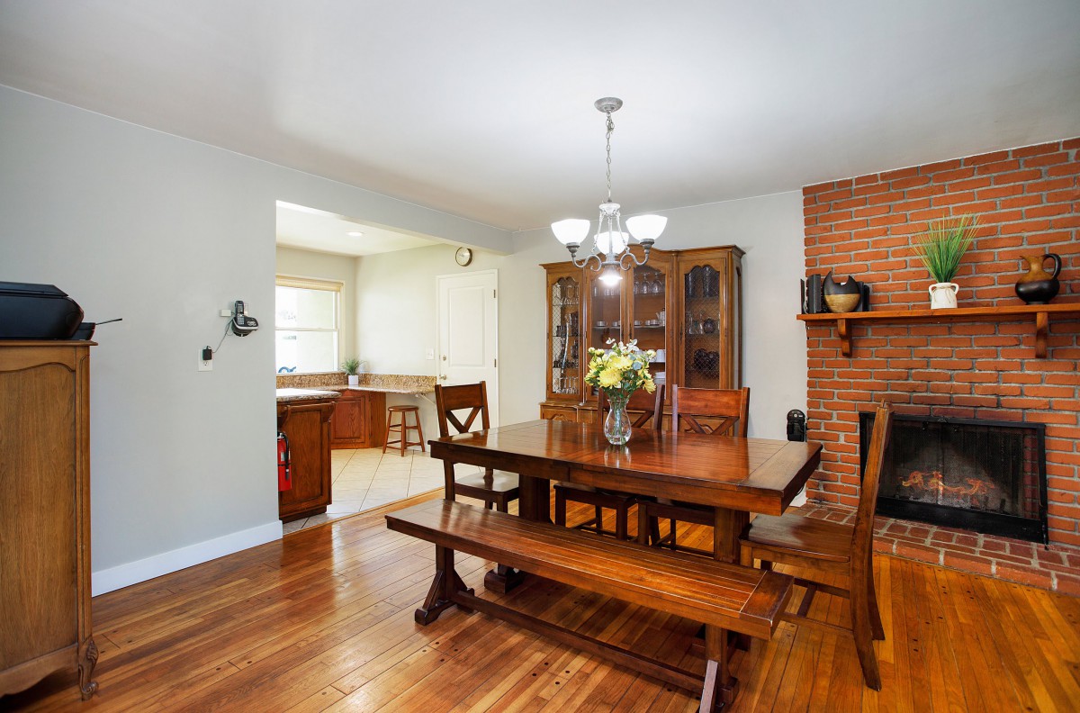 A cozy dining room featuring a wooden table with bench seating, an elegant chandelier, and a brick accent wall, adjacent to a kitchen.