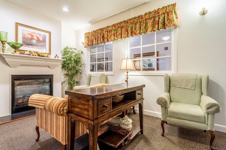 A cozy living room setup featuring a wooden desk, armchairs, a lamp, and a fireplace with decorative curtains and windows.