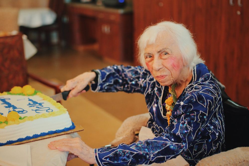 An elderly woman is cutting a cake during a celebration indoors.