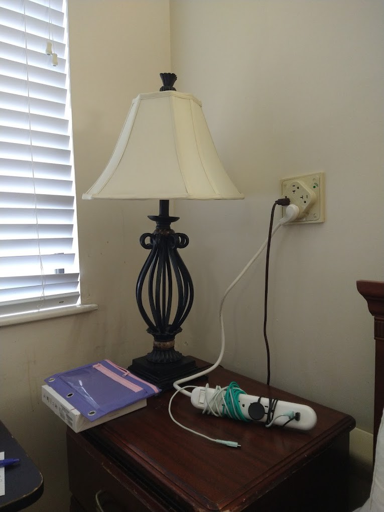 A lamp on a nightstand beside a book and a charging cable plugged into the wall.