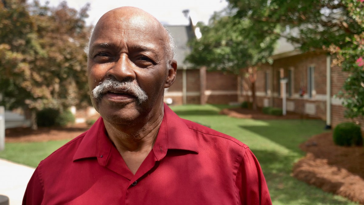 An elderly man wearing a red shirt standing outside in a garden.