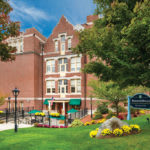 A picturesque brick building set amidst lush greenery and colorful flowers under a clear blue sky.