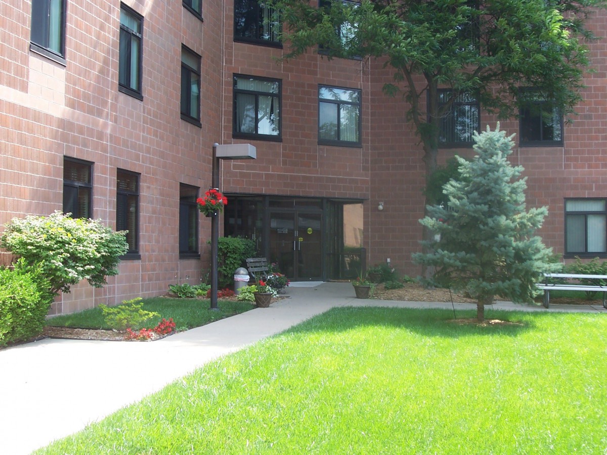An apartment building with a brick exterior and landscaped entrance surrounded by greenery.