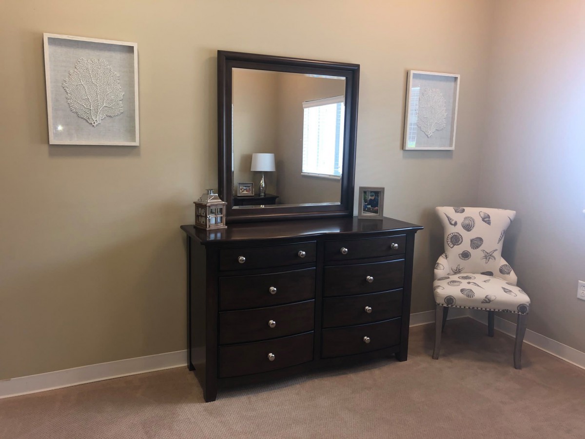 A bedroom dresser with a mirror, two framed pictures on the wall, and an upholstered chair nearby.