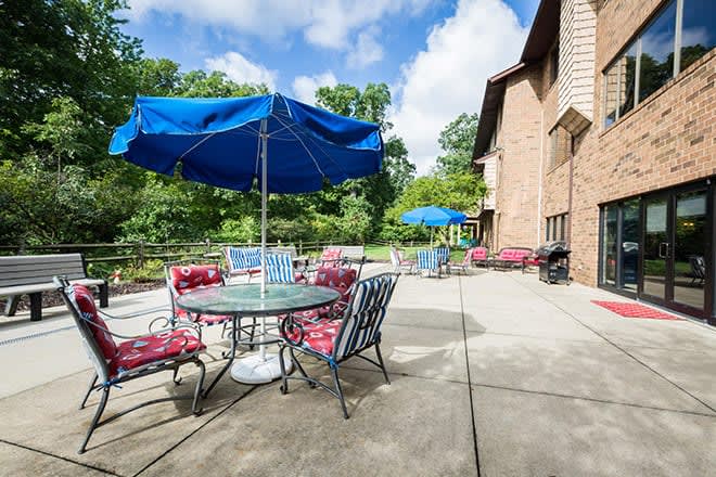 A sunny outdoor patio area with tables, chairs, and blue umbrellas next to a brick building.