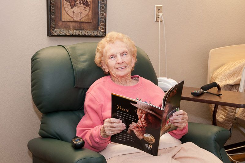An elderly woman sitting comfortably in an armchair, reading a magazine.