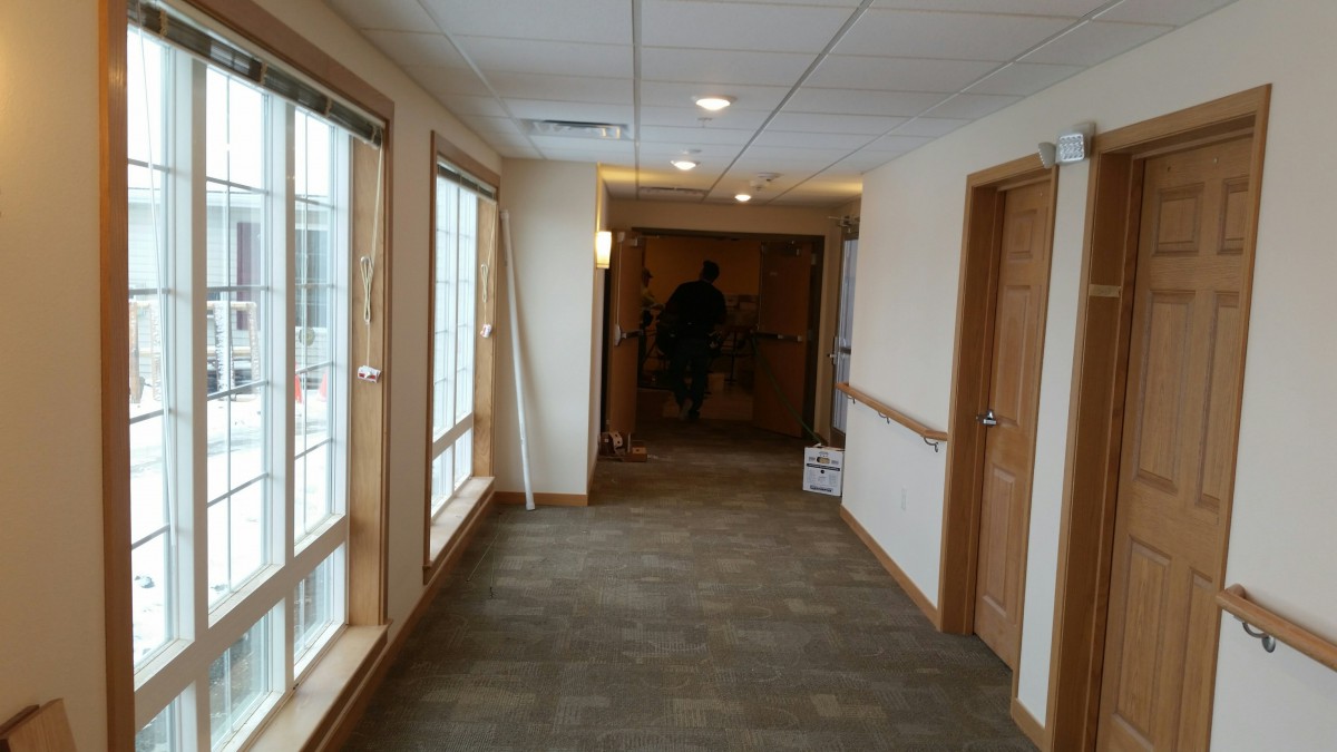 A view of a well-lit hallway with carpet flooring and large windows along one side.