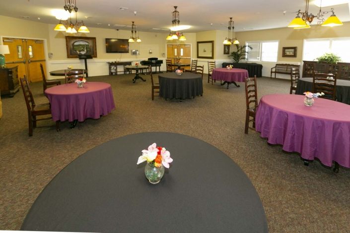 A decorated event room with round tables draped in purple and black tablecloths, featuring flower arrangements.