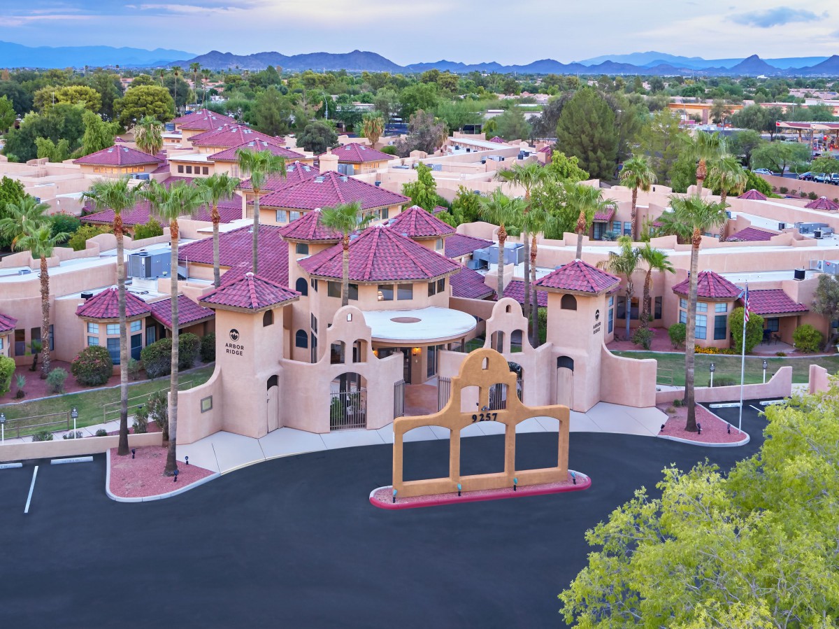 Aerial view of a gated residential community with distinctive architecture and palm trees, set against a backdrop of mountains.