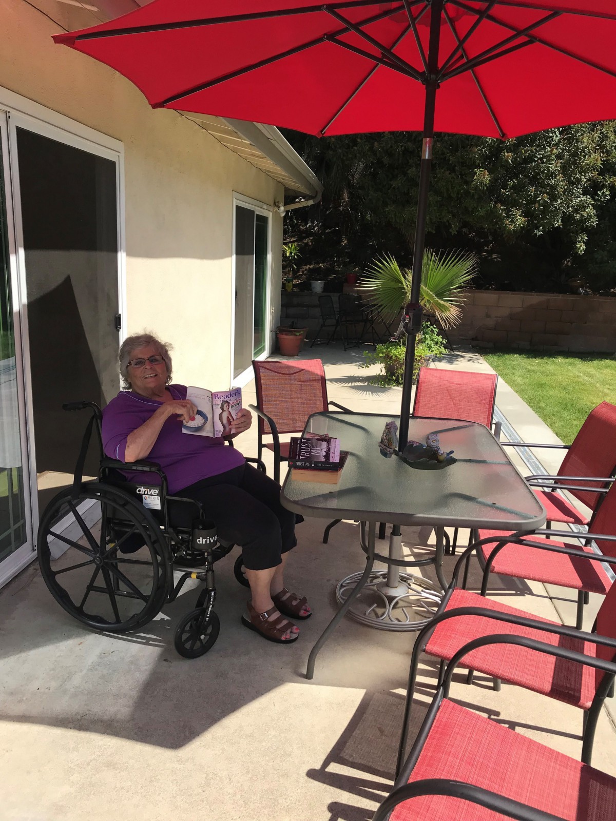 A person in a wheelchair enjoys reading on a sunny patio with red garden furniture and an umbrella.
