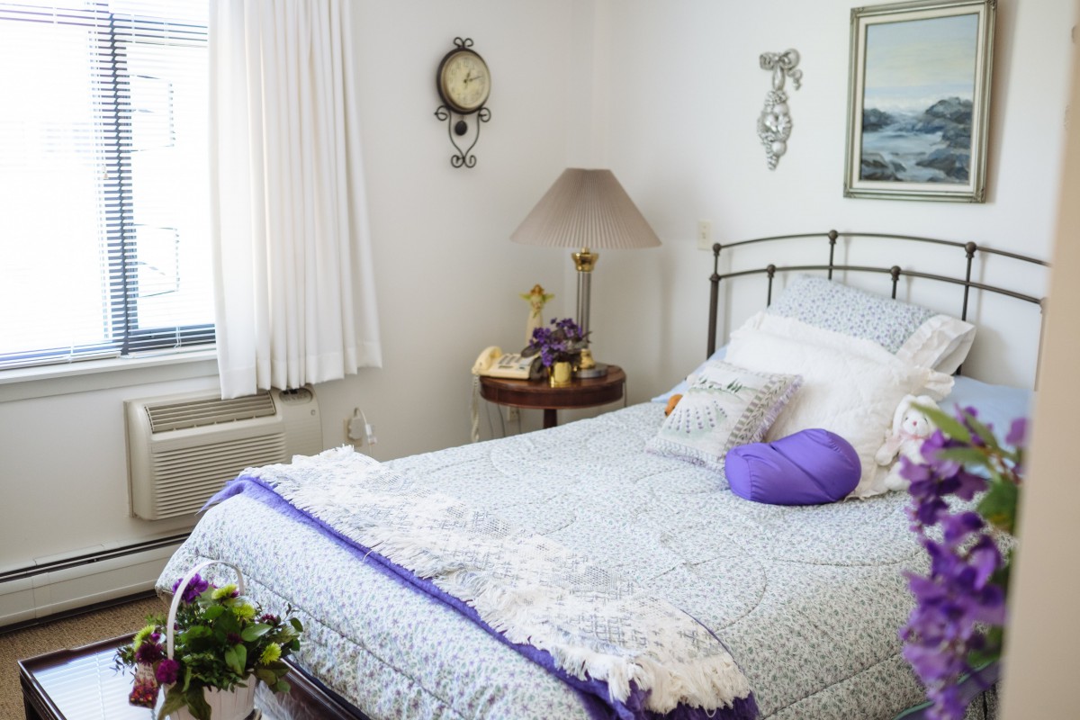 A cozy bedroom with a neatly made bed featuring purple accents, a side table with a lamp and clock, and a window with white curtains.