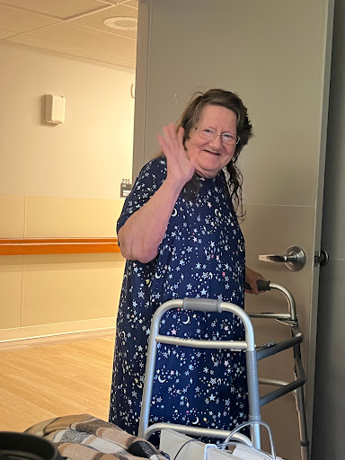 An elderly woman smiles and waves while using a walker in a welcoming indoor environment.