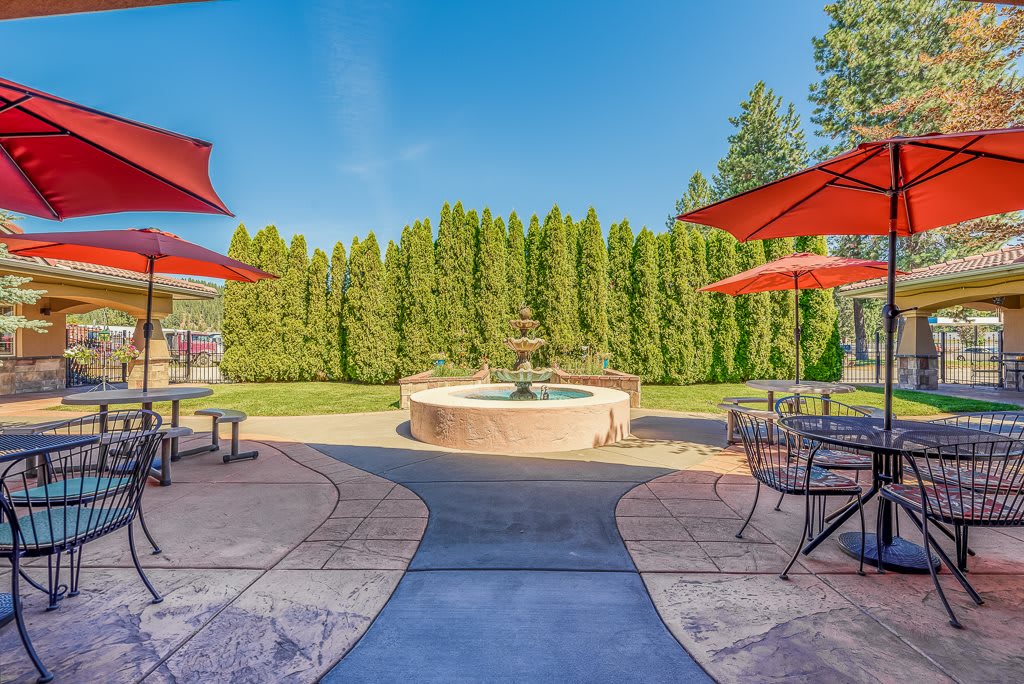 A scenic outdoor patio featuring a central fountain surrounded by tables with red umbrellas and lush greenery.