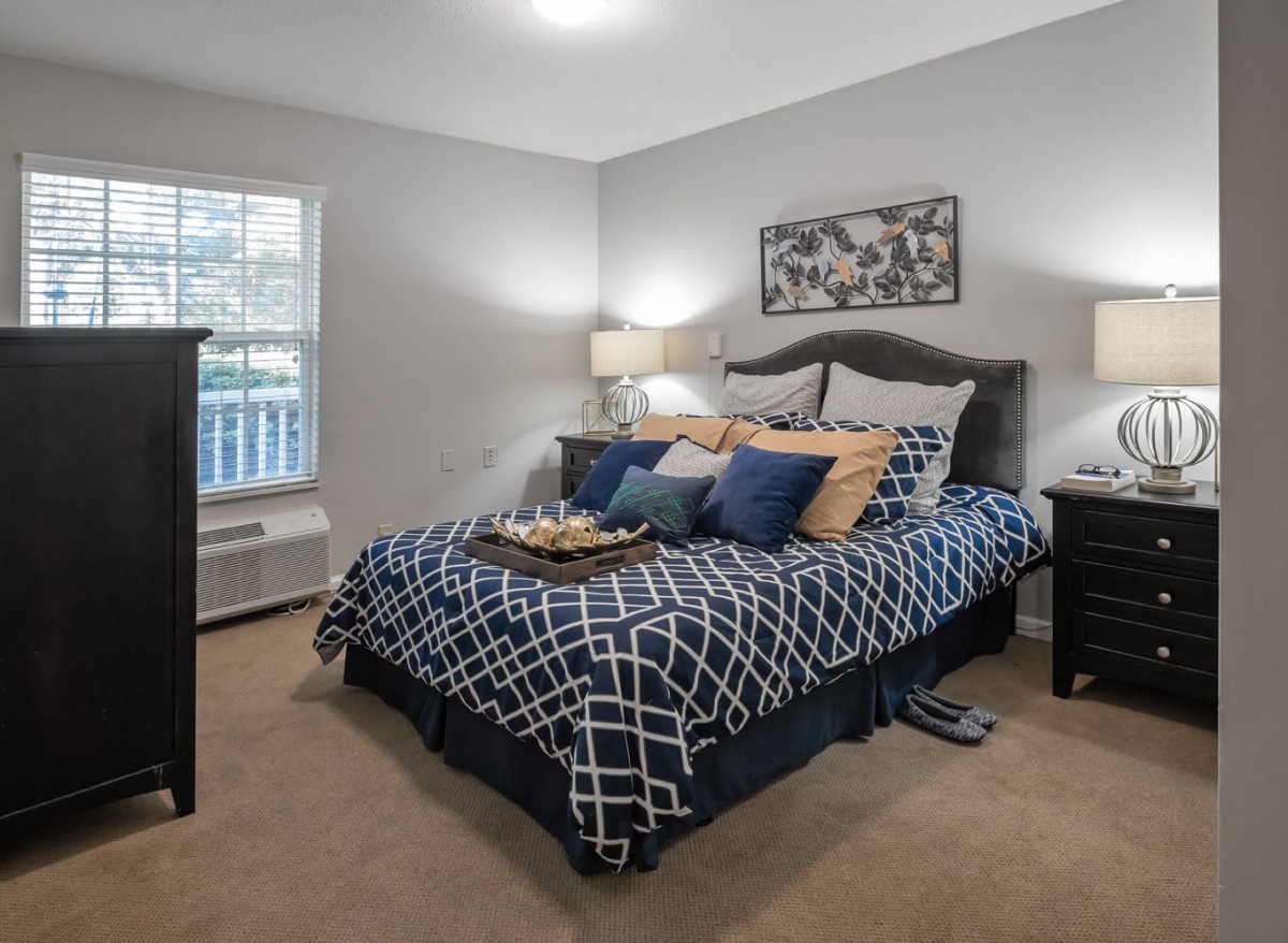A cozy bedroom featuring a neatly made bed with patterned bedding and pillows, flanked by bedside tables with lamps, a window letting in natural light, and carpeted flooring.