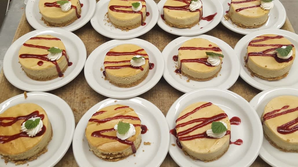 Rows of cheesecakes topped with sauce and garnish on white plates.