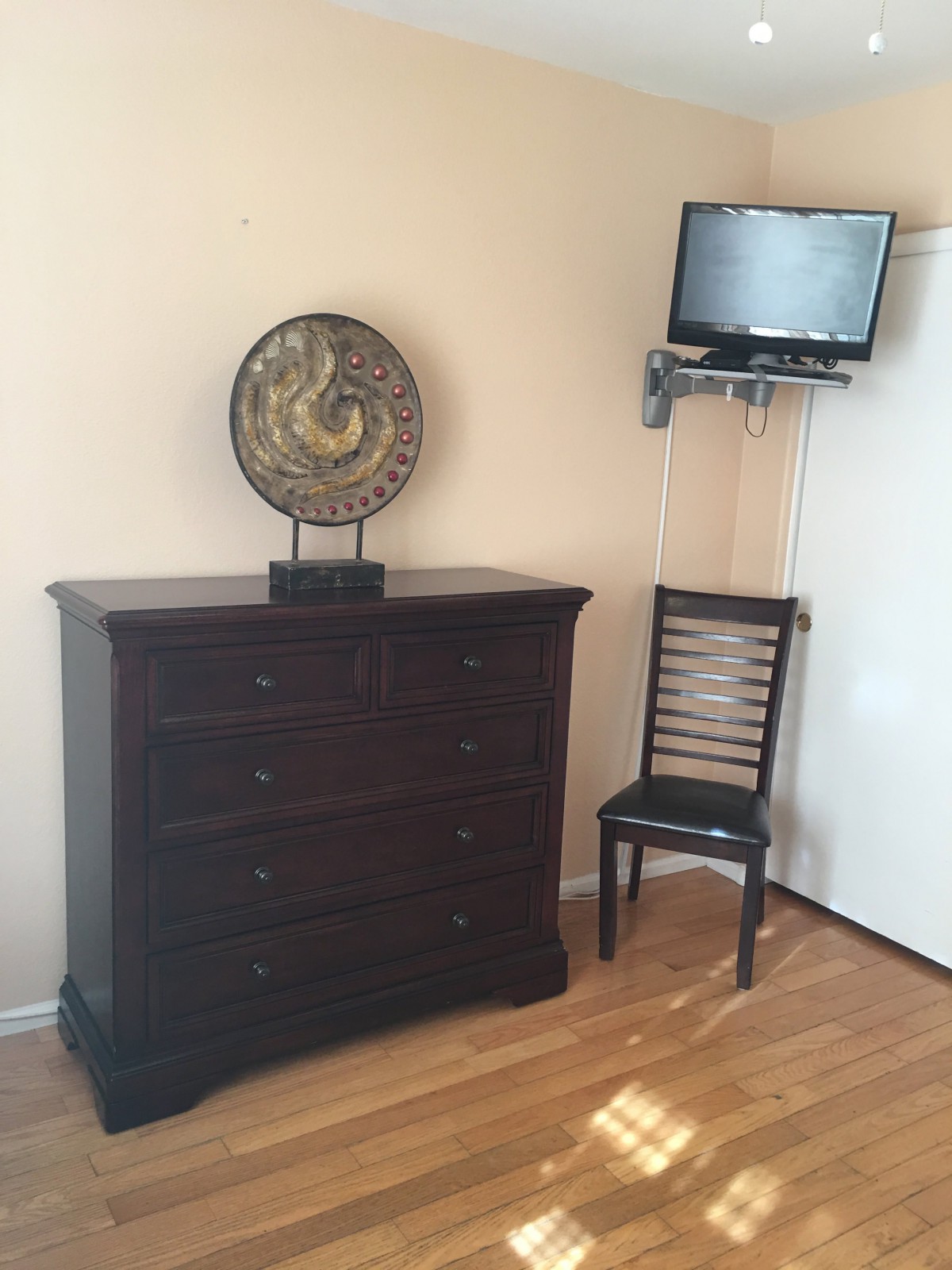A room with a dark wooden dresser, a decorative clock, a chair, and a wall-mounted TV.