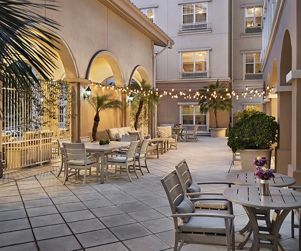 A cozy outdoor patio with string lights, tables, and chairs surrounded by palm trees and building arches.