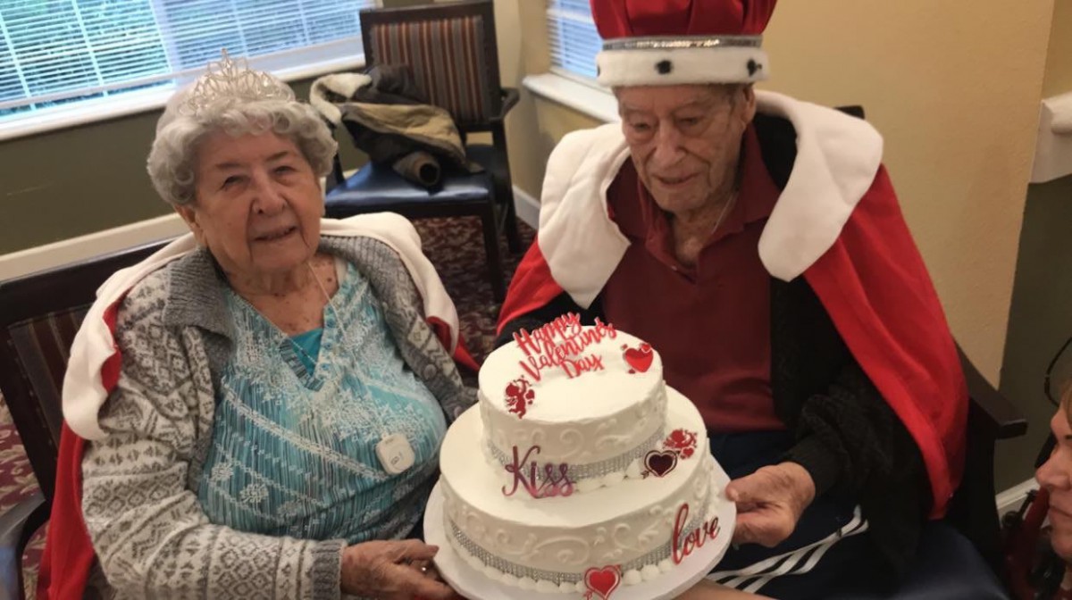 An elderly couple celebrating with a decorated cake, where one person is dressed in a king costume with a crown.
