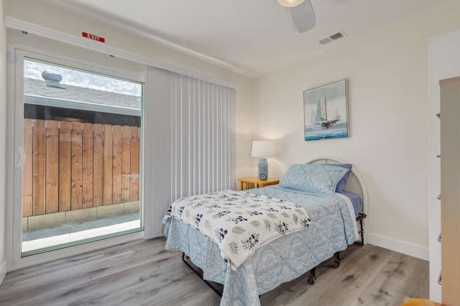 A modern, minimalist bedroom featuring a single bed with blue bedding, a nightstand with a lamp, and a large window letting in natural light.