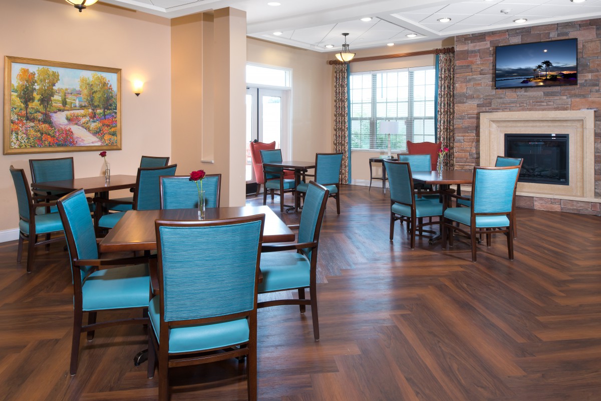 A well-lit dining room with blue chairs and wooden tables, featuring hardwood flooring and large windows.